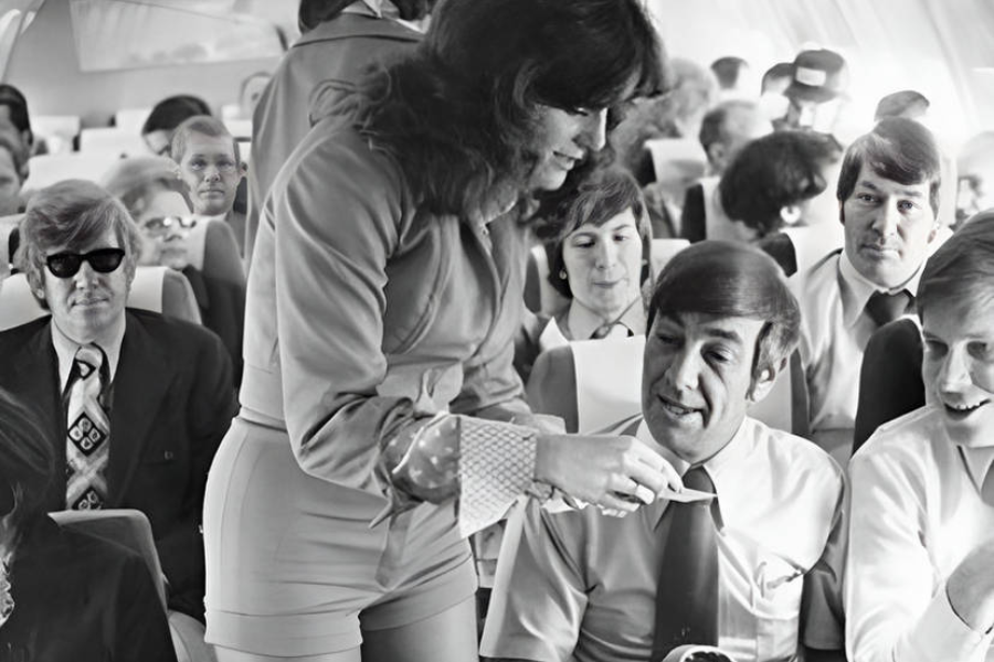 Black-and-white photo of a flight attendant in a fitted uniform serving coffee or tea to a male passenger in a packed airplane cabin. The passengers, dressed in 1970s-style suits and ties, appear relaxed and engaged in conversation.