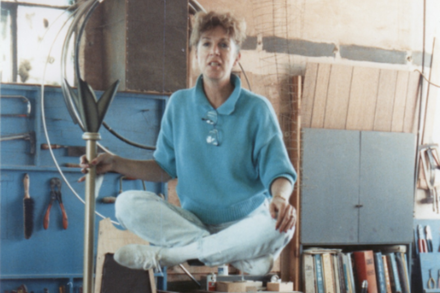 A woman in a blue sweater and light jeans sits cross-legged on a workbench in a workshop, holding a decorative metallic piece resembling a stylized flower or finial. The background includes hand tools on a pegboard, coiled wire, and bookshelves with books and supplies.