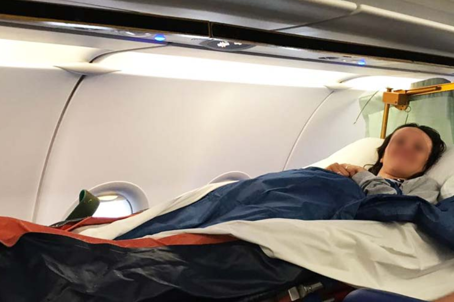 A person lies on a stretcher inside an airplane cabin, covered with a blanket and appearing to rest or sleep. The stretcher is secured along the side of the cabin near the windows and overhead bins.