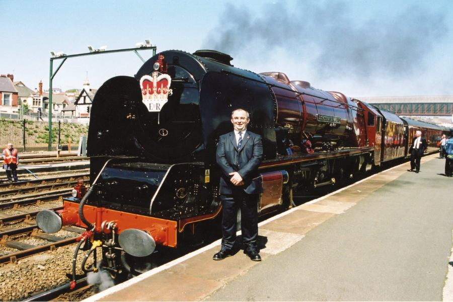 A smiling man in a dark suit stands proudly in front of a glossy, maroon steam locomotive named “The Queen’s Messenger,” stationed on a platform. The front of the engine features a royal emblem with a crown and the letters “E II R,” emitting light steam as it prepares for departure on a sunny day.