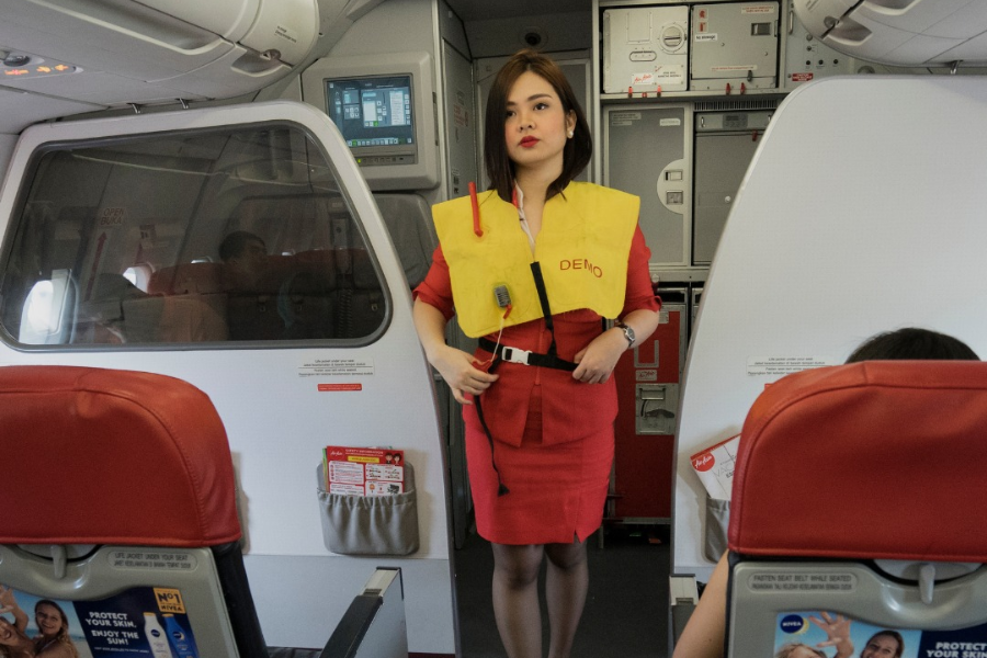 A flight attendant in a red uniform stands at the front of the airplane cabin demonstrating a yellow life vest as part of the safety briefing. The galley and exit door are visible behind her, along with seated passengers.