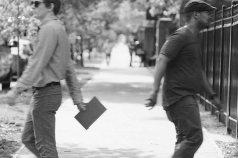 Black and white photo of two men executing a covert handoff on a sidewalk—one casually passing a document folder behind his back to the other as they walk in opposite directions. Trees and parked cars line the street, adding to the subtle tension of the scene.