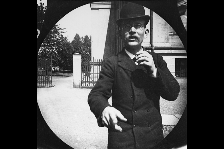 Black and white vintage photo taken through a circular lens, capturing a man in a bowler hat, glasses, and dark suit reaching toward the camera. He stands in front of a gated entrance near a classical building, giving the impression he may have just noticed the hidden camera.