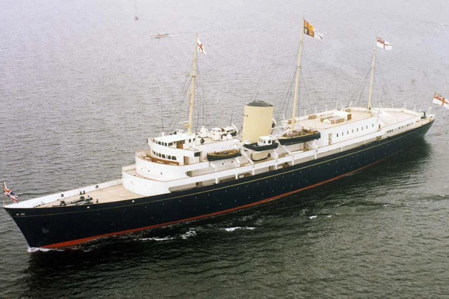 An aerial view of the Royal Yacht Britannia cruising through calm waters. The vessel features a sleek black hull with a white superstructure, cream-colored funnel, and three masts flying the Union Jack and Royal Standard flags. The yacht served as a royal residence at sea and symbol of British diplomacy.