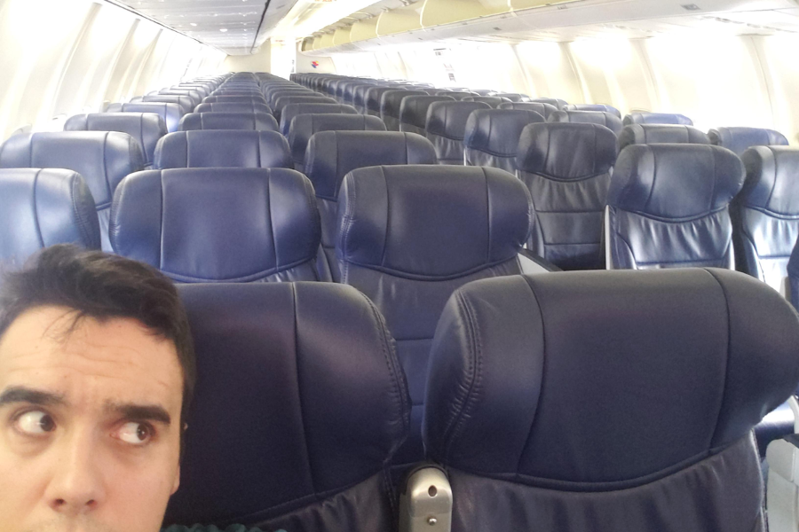 A man takes a selfie on an airplane with rows of empty dark blue leather seats stretching out behind him. He looks off to the side with a surprised or puzzled expression.