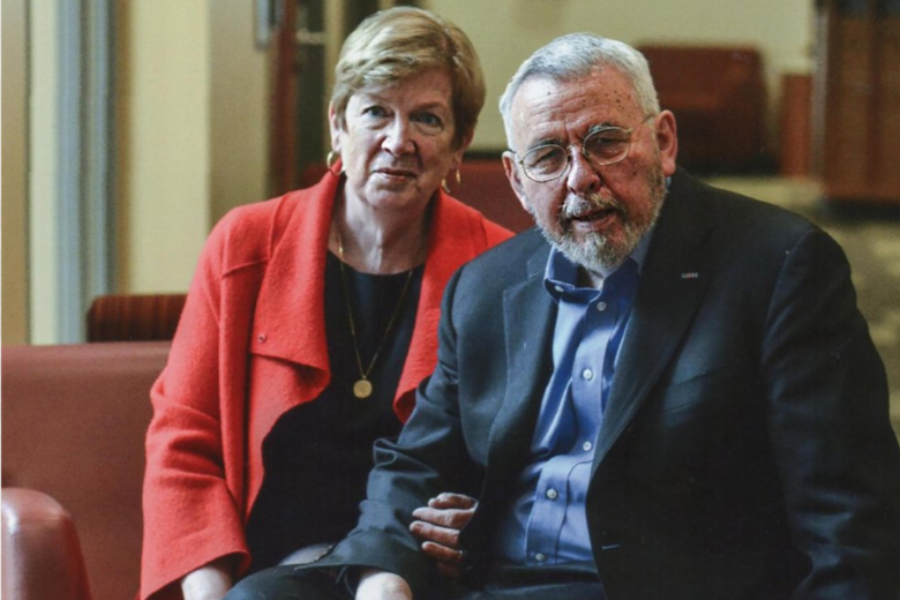 An older couple sits closely together on a red sofa in a warmly lit indoor space. The woman wears a bright red coat and gold pendant, while the man wears glasses and a dark suit jacket over a blue shirt. Both look directly at the camera with a gentle, engaged expression.