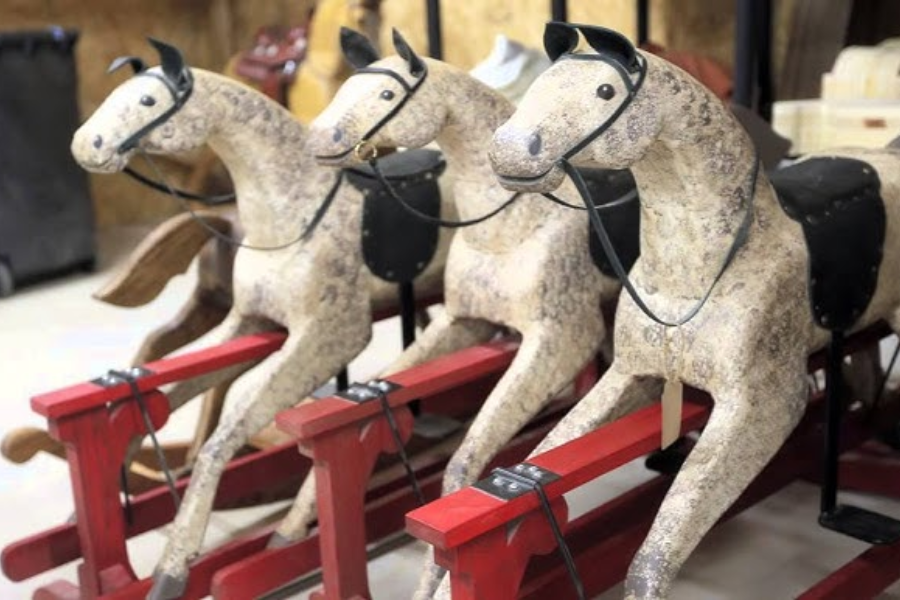 Three vintage-style rocking horses with speckled beige bodies, black leather saddles, and red wooden glider bases are lined up side by side. Each has a bridle and black-painted ears, evoking a nostalgic, handcrafted charm likely intended for children's play or antique decor.