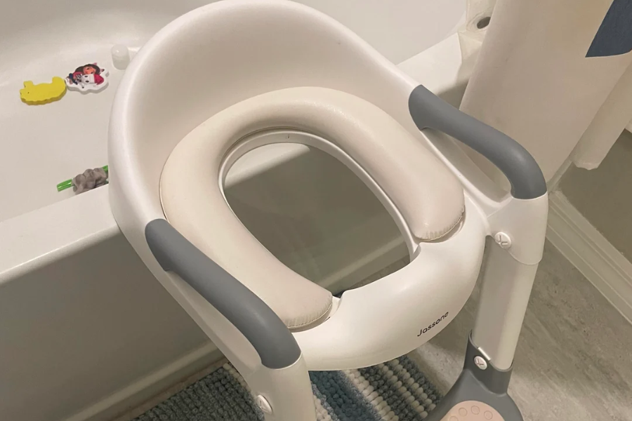 A child’s potty training seat with a white and gray design is positioned in a bathroom next to a bathtub with floating toys. The seat has a soft padded insert and built-in handles for stability.