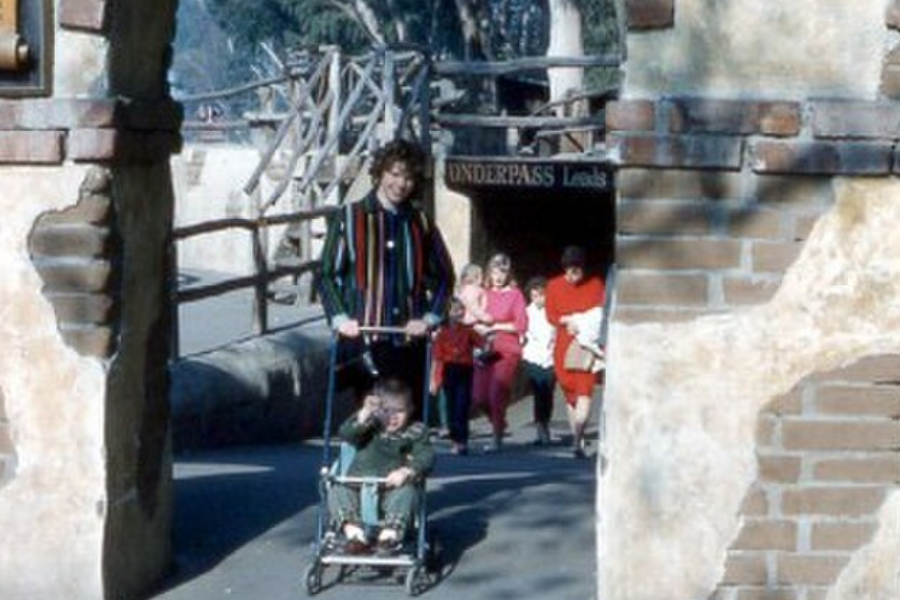 A woman in a colorful striped jacket pushes a young child in a stroller through a rustic stone archway marked “UNDERPASS Leads.” Behind her, a group of adults and children in bright clothing follow, suggesting a family outing or visit to a themed park or zoo.