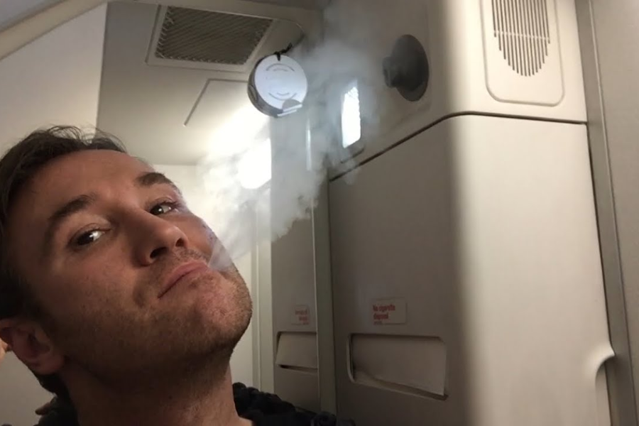 A man on an airplane exhales vapor or smoke near the galley area, with the smoke detector and cabin panels visible in the background. The man appears to be posing for the camera while the vapor rises toward the ceiling.