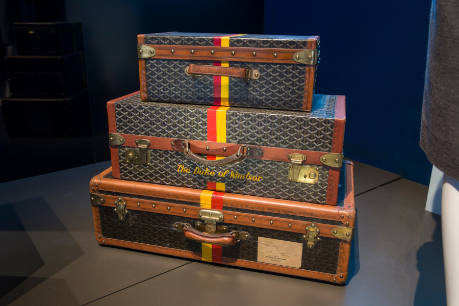 A stacked trio of vintage Goyard suitcases featuring the brand’s iconic chevron pattern, leather trim, and brass hardware. The middle suitcase bears the name "The Duke of Windsor" and all three pieces are adorned with a bold vertical red and yellow stripe, evoking a luxurious, monogrammed travel set once owned by royalty.