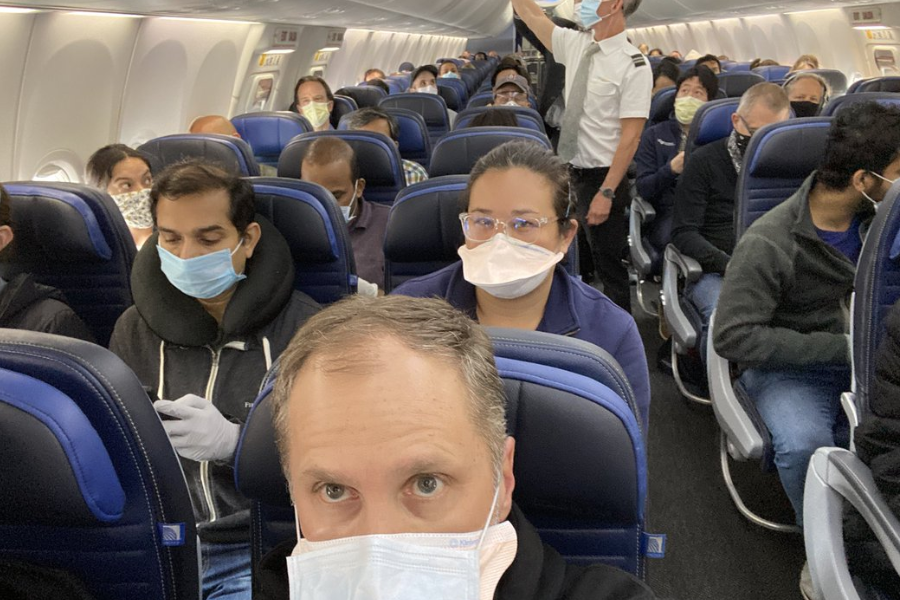Crowded airplane cabin filled with passengers wearing face masks, with a pilot or crew member standing in the aisle. The seats are dark blue with light blue trim, and passengers appear focused or resting.