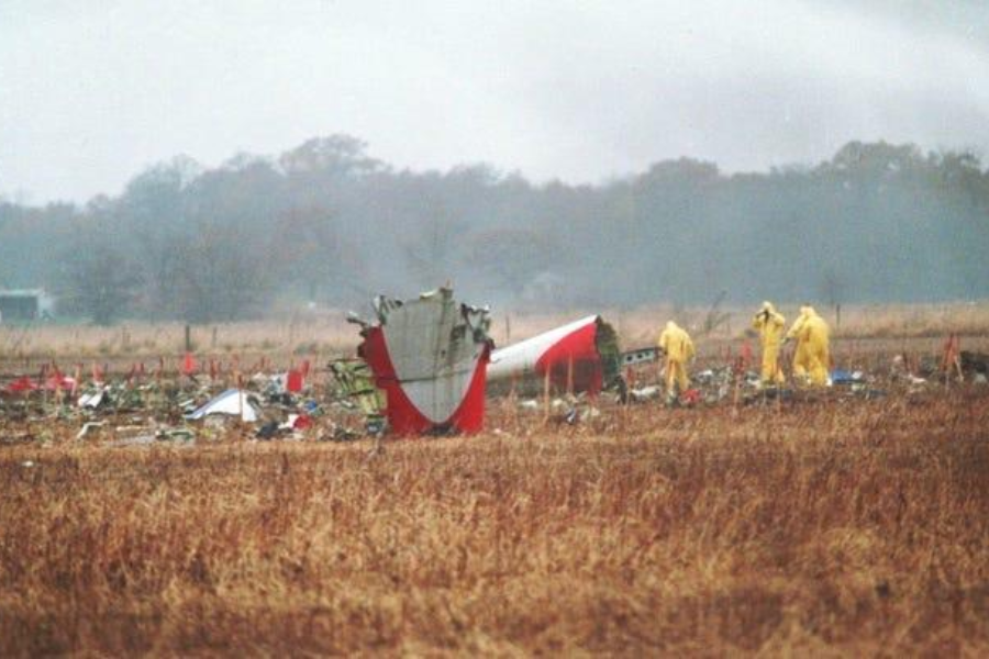 Debris from a crashed airplane scattered across a field, with part of the tail section visibly damaged. Several responders in yellow hazmat suits are seen inspecting the wreckage in the background.