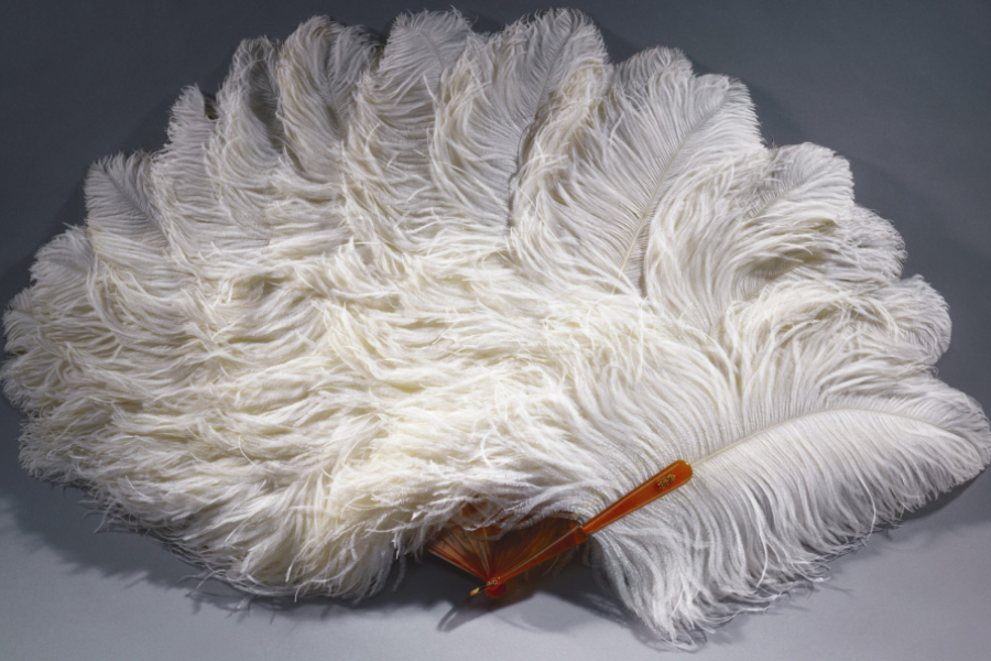 A luxurious hand fan made of large, soft white ostrich feathers arranged in a dramatic semicircle. The feathers are attached to an elegant amber-colored tortoiseshell handle, creating a statement piece likely used in formal occasions or theatrical settings.