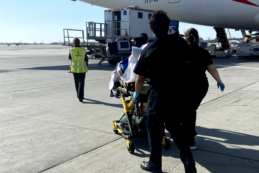 Medical personnel push a patient on a stretcher across an airport tarmac toward a plane, with equipment and monitors visible on the stretcher. A staff member in a yellow safety vest walks ahead, guiding the team.