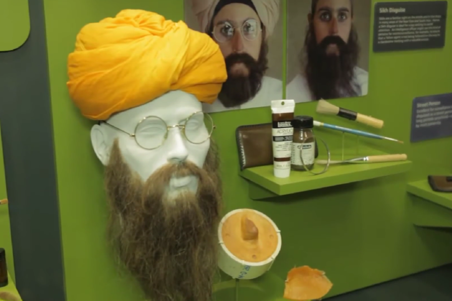 Museum display of a disguise kit replicating a Sikh appearance, featuring a mannequin head with a yellow turban, long beard, and wire-frame glasses. Surrounding items include a prosthetic nose, makeup tools, paints, and reference photos showing the disguise in use.