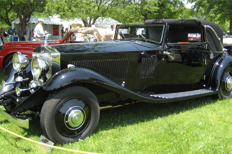 A gleaming black vintage Rolls-Royce convertible with a soft-top roof, displayed at a classic car show on a grassy lawn. The vehicle features a long, elegant body, large chrome headlights, and the iconic Rolls-Royce grille and hood ornament, with reflections of surrounding cars and people on its polished surface.