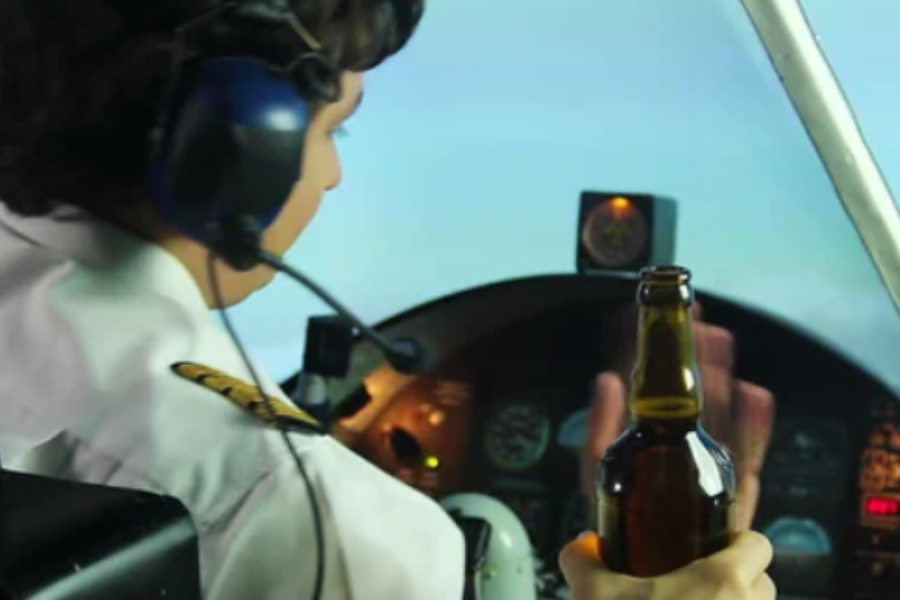 A pilot wearing a white uniform shirt with epaulets and a headset is seated in a cockpit, holding a beer bottle in one hand while the other hand appears to gesture toward the controls. The cockpit instruments are visible in the background.