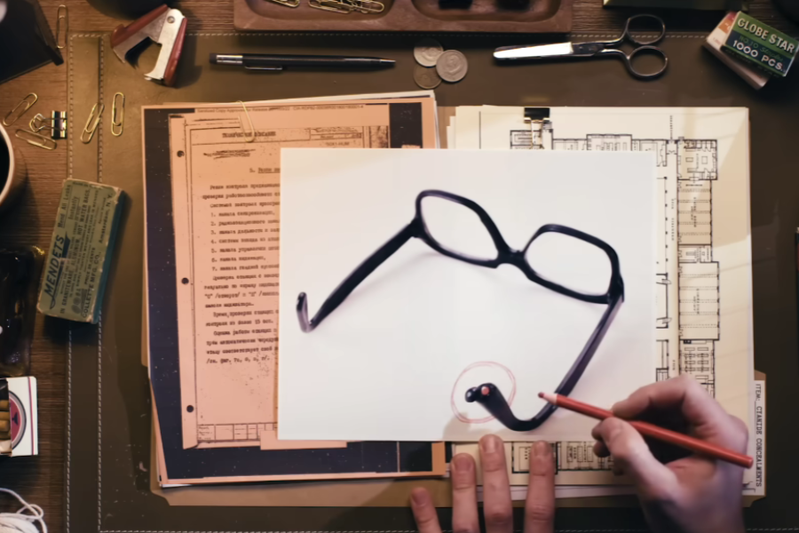 Overhead view of a desk with espionage documents and a photo of black-rimmed glasses containing a hidden device circled in red. A hand holding a red pencil points to the gadget embedded in the temple arm of the glasses, possibly a camera or transmitter.