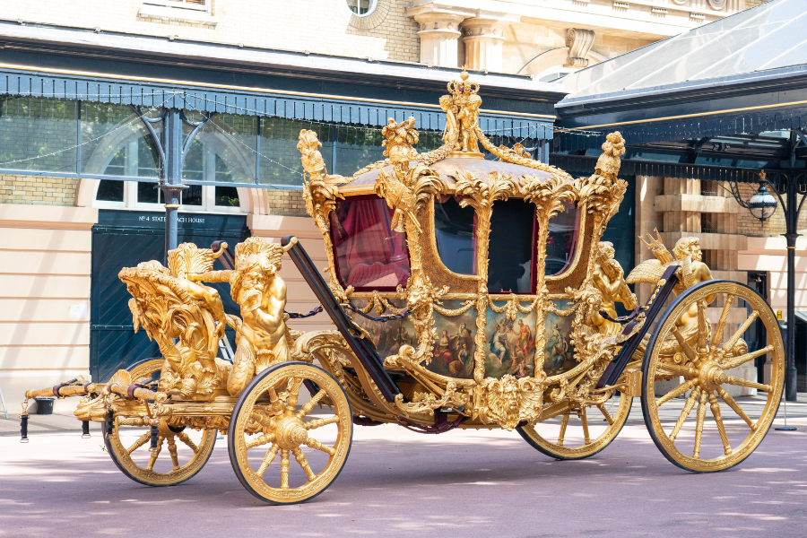 An elaborately decorated royal carriage gilded in gold with intricate carvings, statues, and painted panels, parked outside a stately building. The carriage features large spoked wheels, plush red interior curtains, and ornate sculptures of cherubs and lions, exemplifying opulence and ceremonial grandeur.