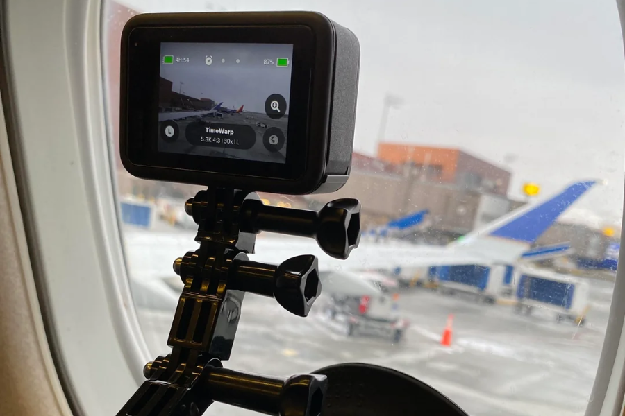 A GoPro camera mounted to the inside of an airplane window records the airport tarmac, with airplanes and ground equipment visible outside. The camera screen shows it is set to TimeWarp mode and capturing in 5.3K resolution.