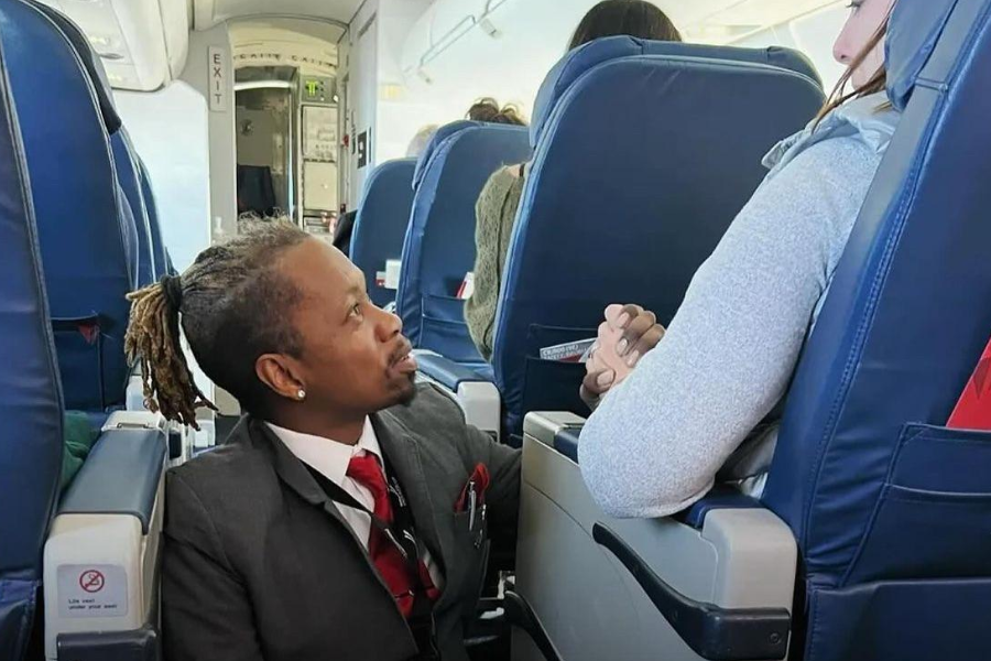 A flight attendant in a suit with a red tie kneels in the airplane aisle, holding hands and speaking gently with a seated passenger. The passenger looks comforted, and the cabin seats and exit door are visible in the background.