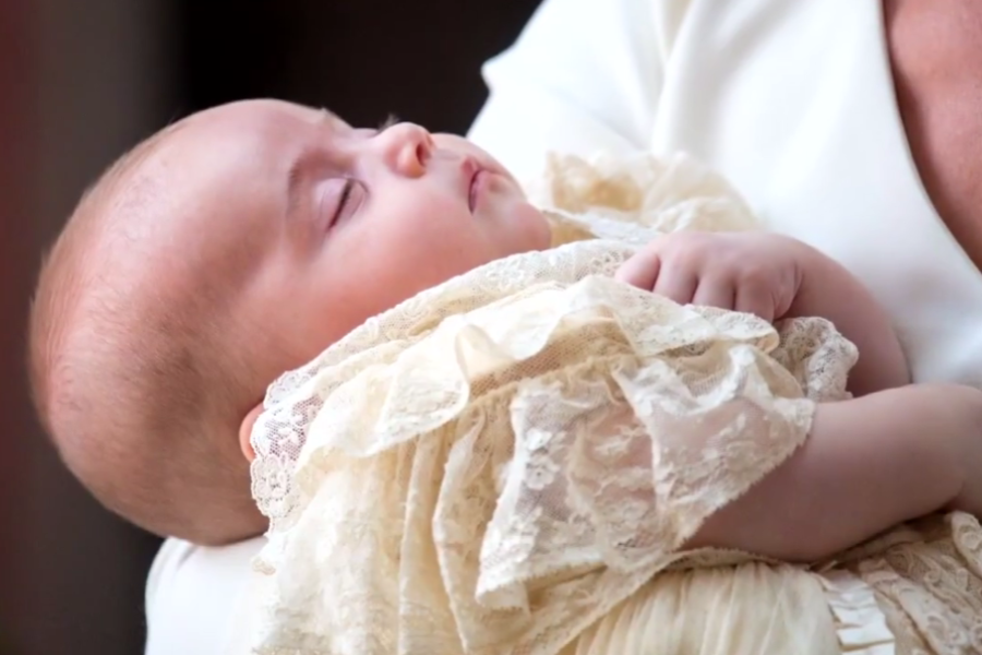 A close-up of a peacefully sleeping infant dressed in an ornate cream-colored lace christening gown, being gently held in an adult’s arms. The traditional gown features ruffled lace trim and delicate embroidery, suggesting a formal or ceremonial occasion such as a royal baptism.