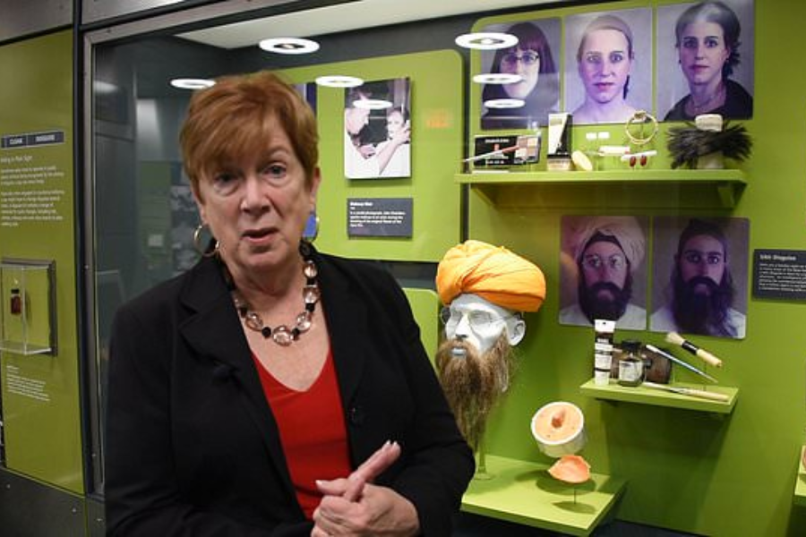A woman stands in front of a spy disguise exhibit at a museum, speaking or gesturing toward the camera. Behind her are mannequins and photographs showing various transformations, including a mannequin head wearing a turban and beard, plus shelves with makeup tools, prosthetics, and disguise materials.