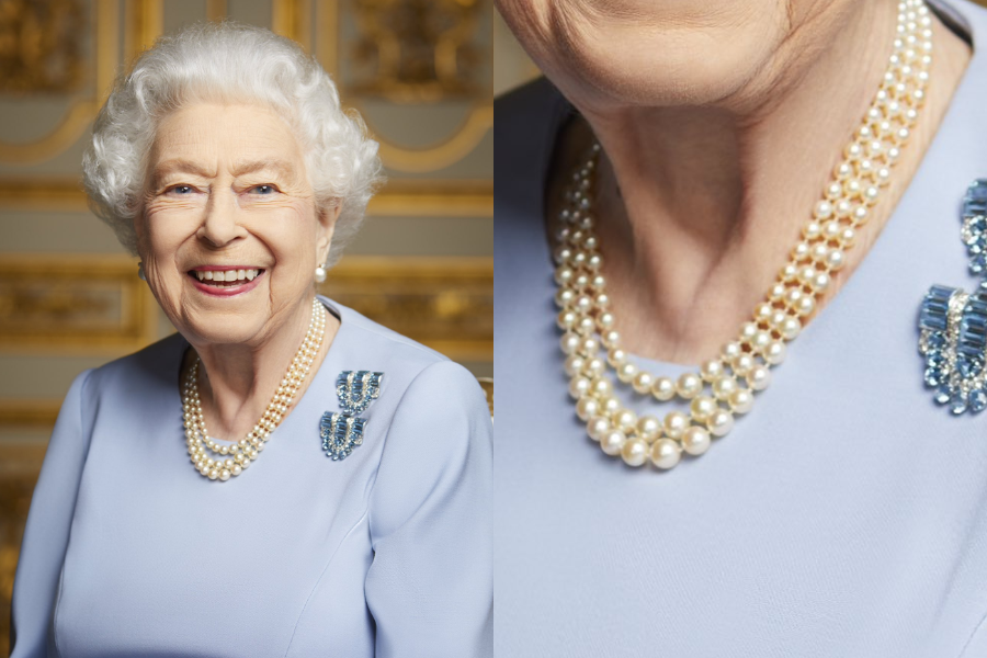 An elderly Queen Elizabeth II smiles warmly while wearing a soft periwinkle blue dress adorned with a triple-strand pearl necklace and a sparkling aquamarine brooch set. A close-up on the right highlights the creamy luster of the pearls and the detailed design of the brooches pinned to her shoulder.