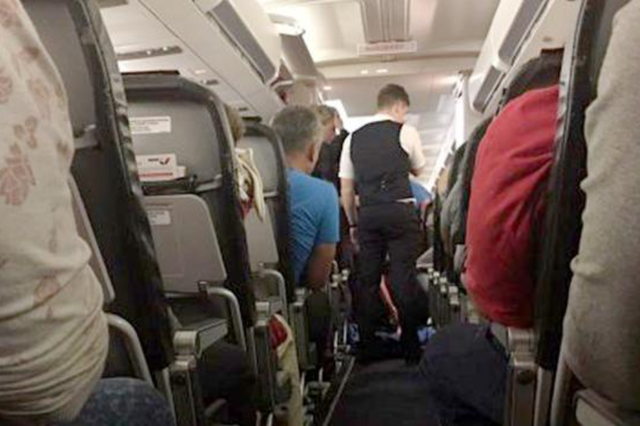 View down the aisle of an airplane where a flight attendant in a black vest appears to be assisting with a situation near the front of the cabin. Seated passengers look on, and the cabin is filled with standard seats and tray tables.