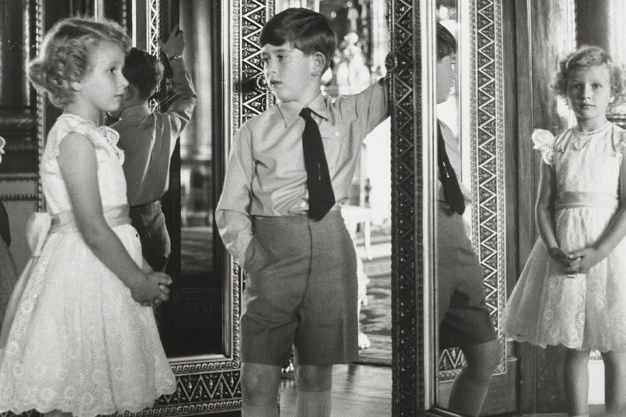 A black-and-white photo of two young children standing in an ornate mirrored room. The girl wears a delicate lace dress with a ribbon sash, while the boy wears a collared shirt, tie, and high-waisted shorts. Their reflections appear multiple times in the large decorative mirrors surrounding them, creating a layered and nostalgic composition.