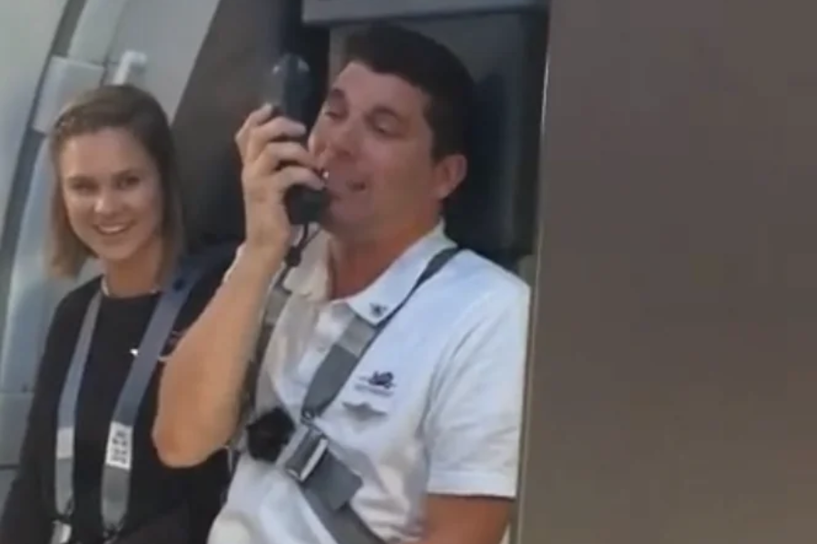 Two flight attendants seated near an airplane door, with one smiling and the other speaking into the cabin intercom handset. Both are wearing seat harnesses and appear to be engaging with passengers.