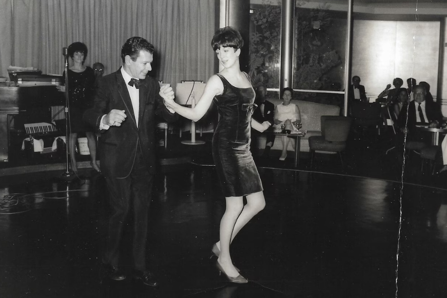 Black and white photo of a man in a tuxedo and a woman in a sleeveless velvet dress dancing on a stage in front of an audience at what appears to be a lounge or nightclub. Seated patrons in formal attire watch the performance, while a woman stands near a microphone and a piano in the background.