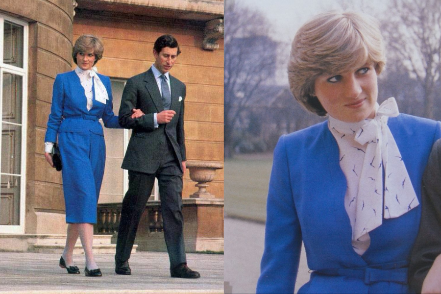 A woman in a vibrant cobalt blue suit with a white silk blouse featuring a subtle bird print walks arm-in-arm with a man in a dark suit outside a grand stone building. The close-up highlights her softly feathered hairstyle and the delicate bow tied at her neck, adding a refined touch to her polished look.
