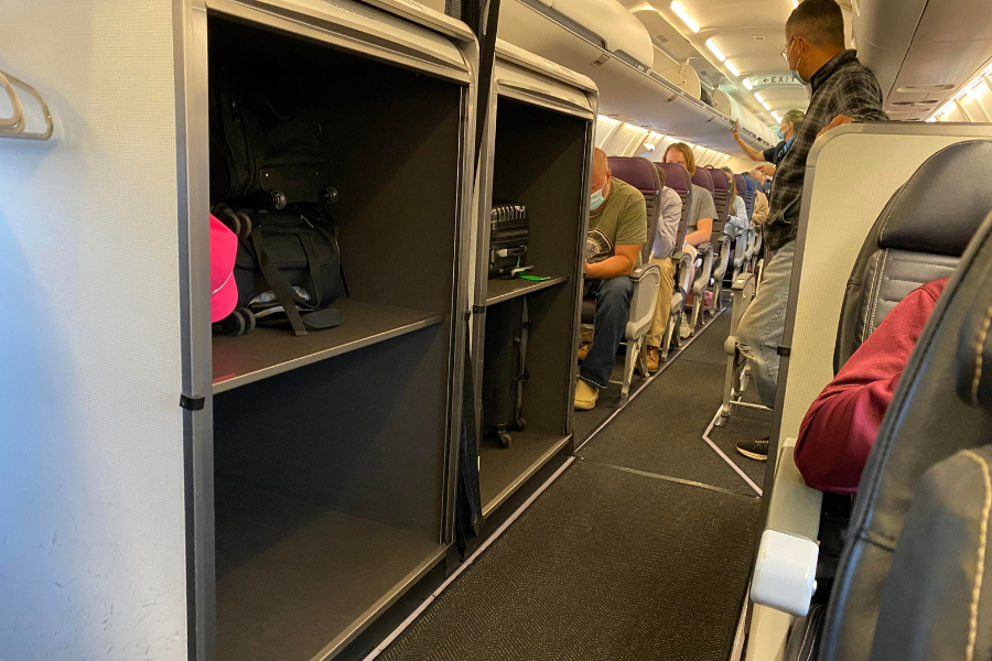 Interior of an airplane showing an open luggage storage area near the front of the cabin, with a few bags and a bright pink item on the shelves. Passengers are seated and wearing masks, while one man stands in the aisle.