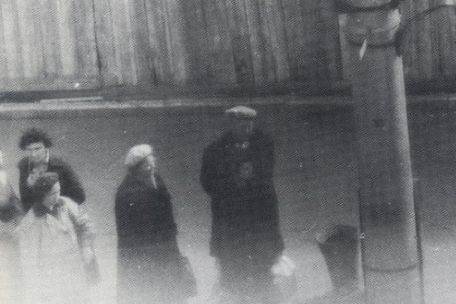 Grainy black and white photo showing a group of five people walking on a sidewalk near a tall utility pole. Most are wearing coats and hats, suggesting a cold day, with a wooden fence lining the background.