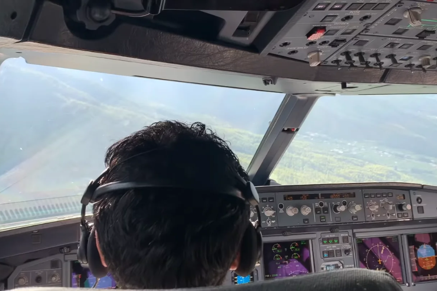 View from behind a pilot wearing headphones in the cockpit of an aircraft, with cockpit controls and displays visible. The plane appears to be banking over a landscape of hills and greenery seen through the windshield.