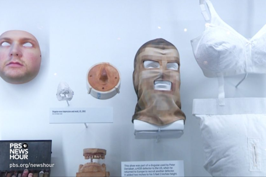 Display case containing various espionage disguises and props, including a hyper-realistic face mask, a sculpted nose impression, a pixelated balaclava-style mask, and a white bra with padding. The exhibit is part of a PBS NewsHour segment on spy tactics, featuring objects once used by intelligence operatives.