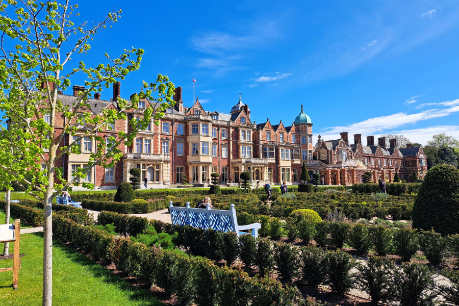 A stately red-brick mansion with ornate chimneys and a green copper dome rises behind manicured gardens at Sandringham House in Norfolk, England. Visitors stroll along gravel paths and sit on white benches amid neatly trimmed hedges, all under a clear blue sky.