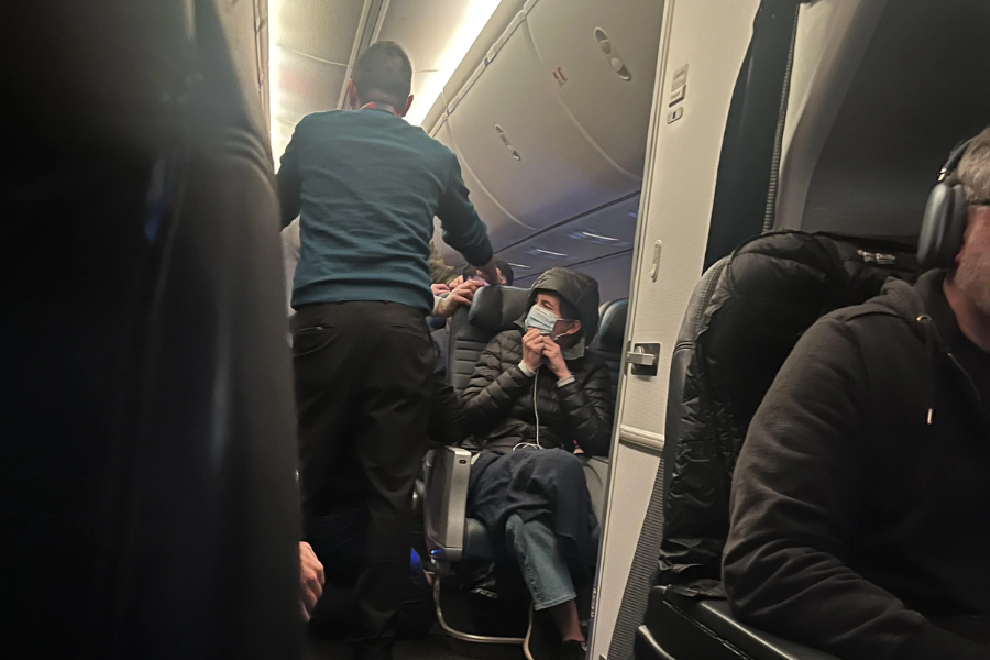 Airplane cabin scene showing a man in a blue sweater standing in the aisle near the front, while a seated passenger in a hooded jacket and face mask looks on. Other passengers are seated nearby, some wearing headphones, in a dimly lit cabin.
