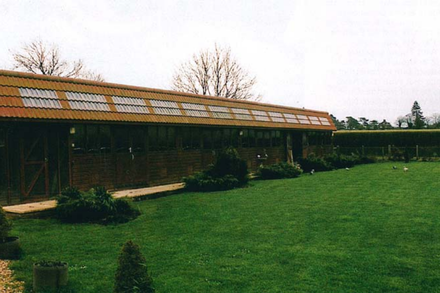 A long, low wooden outbuilding with a tiled roof and rows of skylights stands beside a neatly maintained grassy lawn. Surrounded by small shrubs and trees, the structure appears to be a stable or aviary, set in a quiet, fenced rural landscape.