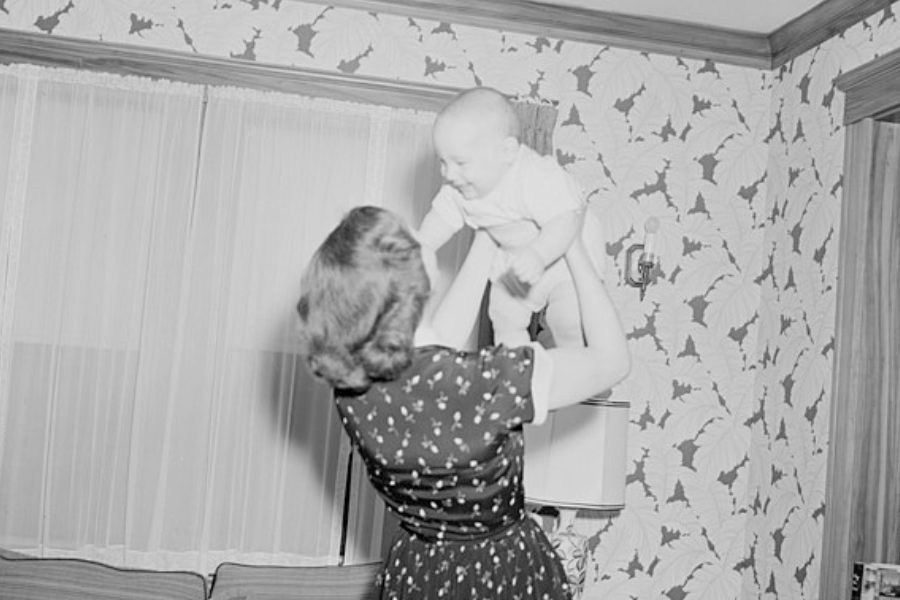 Black and white photo of a woman in a patterned dress lifting a smiling baby into the air inside a home with leaf-patterned wallpaper and lace-curtained windows. The baby looks joyful, and the moment captures a warm, playful interaction.