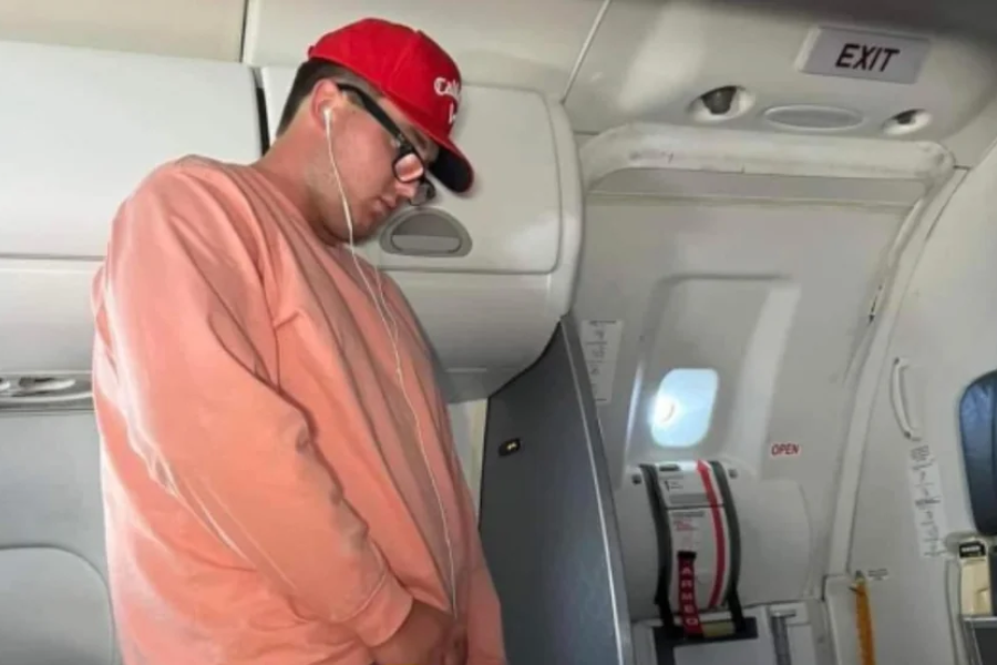 A man wearing a red cap, glasses, and a pink sweatshirt stands near the exit door of an airplane, appearing to urinate or simulate urination. He has earphones in and is facing downward, with an overhead bin and emergency exit signage visible.