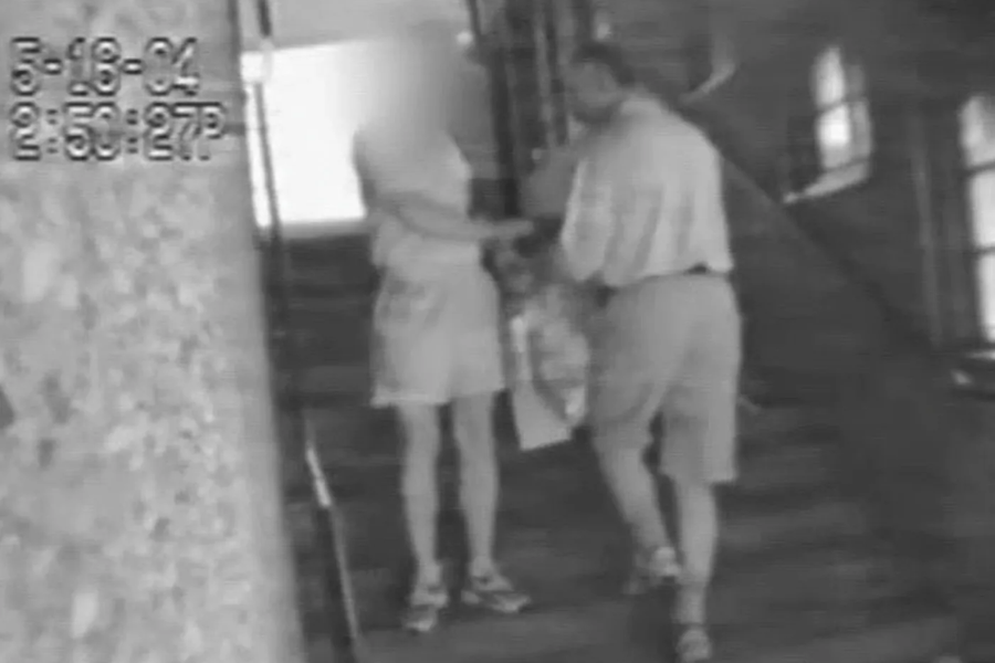 Grainy black and white surveillance footage showing two people on a staircase, one handing a bag to the other. The timestamp reads "5-19-04 2:50:27 PM," and the face of the person on the left is blurred for anonymity.