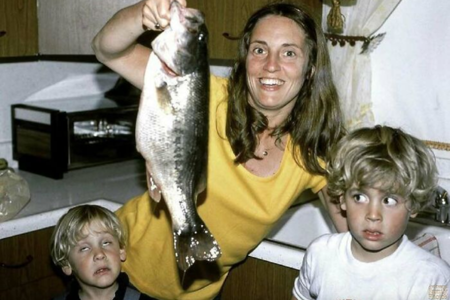 A woman proudly holds up a large fish in a kitchen, while two young boys beside her make exaggerated, disgusted faces.