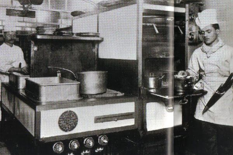 Black and white photo of chefs working in the Titanic's kitchen, showing industrial stoves, large pots, and a chef preparing food beside a mounted oven.