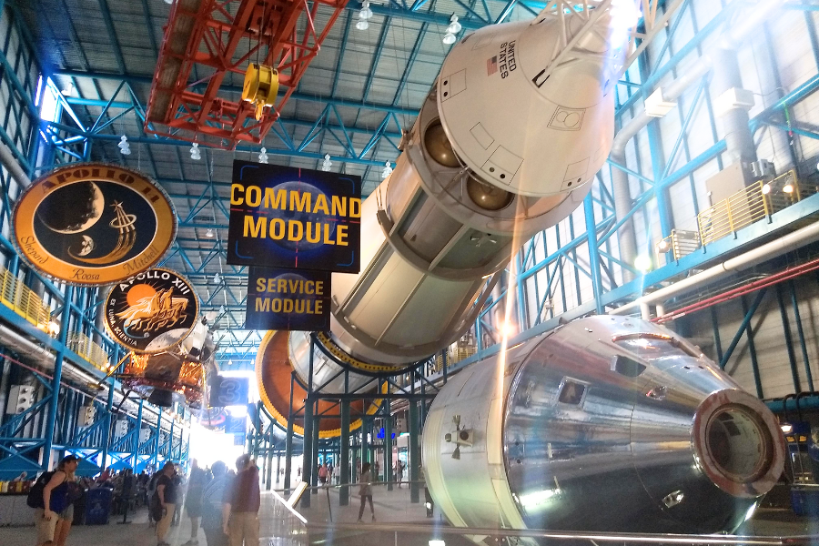 Visitors explore a NASA exhibit hall showcasing the Apollo spacecraft’s command and service modules, suspended in dramatic lighting with signage and mission patches.
