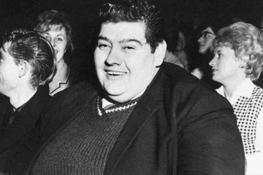 Black and white photo of a smiling man in a dark suit and sweater, seated in a crowded audience with people around him chatting and laughing.