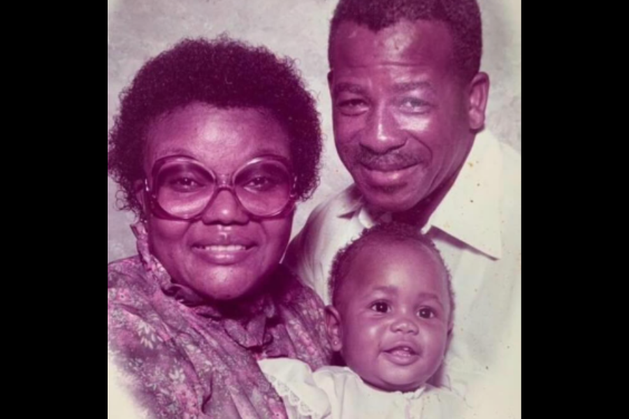 A vintage portrait of a smiling couple holding a baby, with the woman wearing large glasses and a floral dress, and the man in a light-colored collared shirt. The photo has a warm, nostalgic tint.
