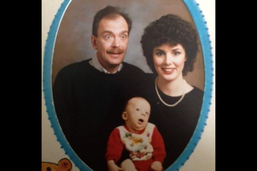 A couple poses with their baby for a professional portrait, though the baby looks hilariously unamused and appears mid-yawn or scream.